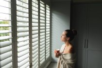 Vista lateral da mulher caucasiana com caneca de café olhando através da janela em casa — Fotografia de Stock
