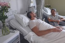 Вид сбоку на пациентов мужского пола, взаимодействующих друг с другом в палате больницы. Белый пациент лежит в постели, в то время как пациент смешанной расы сидит в инвалидном кресле . — стоковое фото