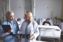 Frontansicht diverser Ärztinnen, die auf der Station im Krankenhaus über Röntgenberichte diskutieren. Im Hintergrund interagieren verschiedene Ärzte mit ihren Patienten. — Stockfoto