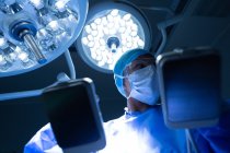 Хирург смешанной расы с низким углом обзора удерживает дефибриллятор, глядя в операционную в больнице — стоковое фото