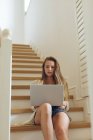 Vista frontal de la hermosa mujer caucásica utilizando el ordenador portátil en las escaleras en casa - foto de stock