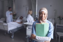 Vista frontal de la doctora de raza mixta en hiyab de pie con expedientes médicos en la sala del hospital. En el fondo médicos caucásicos revisando a los pacientes . - foto de stock