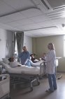 Вид збоку різних лікарів взаємодіючих з пацієнтами у приході в лікарні. У фоновому режимі Кавказький людина тримає руку азіатській жінці, яка лежала в ліжку в лікарні. — стокове фото