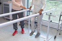 Low section of female physiotherapist helping mixed-race female patient walk with parallel bars in the hospital — Stock Photo