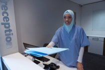 Vista frontal de la cirujana de raza mixta en hijab de pie con archivo de informe médico en recepción en el hospital - foto de stock