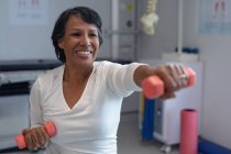 Vista frontal de una paciente de raza mixta que hace ejercicio con pesas anaranjadas en el hospital - foto de stock