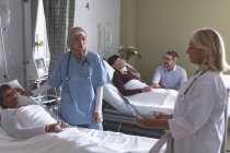 Vue de face de divers médecins féminins qui interagissent avec des patientes à l'hôpital. Dans le fond l'homme caucasien tient la main de la femme asiatique qui est couchée dans le lit à l'hôpital . — Photo de stock