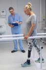 Side view of Caucasian male physiotherapist writing on clipboard and Caucasian female amputee patient walk with parallel bars in the hospital — Stock Photo