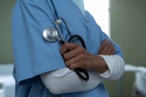 Mittelteil einer Ärztin, die mit verschränkten Armen auf der Station im Krankenhaus steht, während sie Stethoskop in der Hand hält — Stockfoto