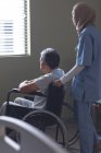 Visão lateral da médica mista em hijab em pé com o paciente do sexo masculino em cadeira de rodas no hospital . — Fotografia de Stock