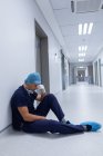 Vista laterale del bel chirurgo caucasico teso con mano sul viso seduto nel corridoio dell'ospedale. Il chirurgo indossa maschera chirurgica, berretto chirurgico, abito, e guanti chirurgici . — Foto stock