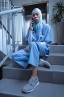Vista frontal de la enfermera de raza mixta en hijab hablando por teléfono móvil mientras está sentada en las escaleras en el hospital - foto de stock