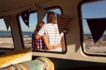 Vista trasera de pareja caucásica con los brazos alrededor de pie juntos cerca de autocaravana en la playa - foto de stock