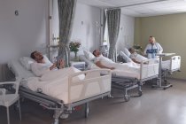 Огляд різноманітного пацієнта чоловічої статі, який використовує цифровий планшет або сон, а жінка-лікар перевіряє пацієнта чоловічої статі в лікарні — стокове фото