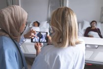 Vista trasera de diversas doctoras discutiendo sobre el informe de rayos X en tableta digital en la sala del hospital. Diversas pacientes femeninas están durmiendo en la cama en el fondo . - foto de stock