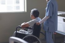 Vue latérale du médecin féminin métis debout avec un patient masculin métis âgé en fauteuil roulant à l'hôpital — Photo de stock