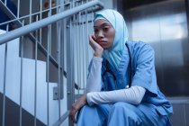 Обзор хирурга смешанной расы в хиджабе, сидящего на лестнице в больнице — стоковое фото