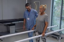 Зовнішній вигляд Кавказького чоловічого фізіотерапевта допомагає кавказька жінка ампутованими кінцівками, ходити з паралельними барами в лікарні — стокове фото
