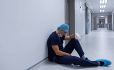Vista lateral do belo cirurgião masculino caucasiano tenso com luvas cirúrgicas sentado no corredor do hospital — Fotografia de Stock