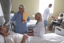 Вид спереди на разнообразных женщин-врачей, взаимодействующих с кавказским пациентом в палате больницы. На заднем плане кавказский врач пожимает руку инвалиду старшему пациенту смешанной расы в инвалидном кресле . — стоковое фото