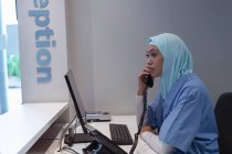 Vista lateral de cirurgiã mestiça em hijab conversando via telefone fixo na recepção do hospital — Fotografia de Stock