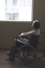 Vista lateral del paciente discapacitado de raza mixta en silla de ruedas mirando a través de la ventana en la sala del hospital - foto de stock