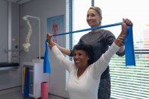 Side view of Caucasian female physiotherapist giving physical therapy with resistance band to mixed race female patient in the hospital — Stock Photo