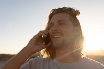 Vista frontal de hombre caucásico joven feliz hablando en el teléfono móvil en la playa - foto de stock
