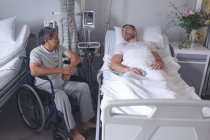 Vue de face de divers patients masculins interagissant les uns avec les autres dans le service de l'hôpital. Patient masculin caucasien allongé au lit tandis que le patient métis est assis en fauteuil roulant . — Photo de stock
