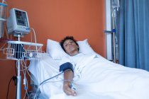 Vista frontal de una paciente de raza mixta tumbada en la cama en la sala del hospital - foto de stock