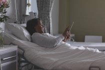 Vue latérale d'une jolie patiente métisse utilisant une tablette numérique à l'hôpital . — Photo de stock
