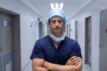 Portrait d'un beau jeune chirurgien caucasien debout les bras croisés dans le couloir de l'hôpital. L'homme porte un masque chirurgical, une robe et un bonnet . — Photo de stock