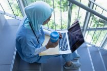 High view of mixed race female doctor in hijab using laptop while having cup of coffee on staircase in hospital — Stock Photo