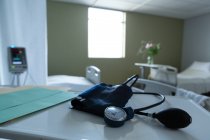 Крупный план измерителя артериального давления и медицинских карт на столе с пустыми кроватями и монитором на заднем плане в палате больницы — стоковое фото