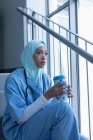 Вид збоку вдумлива змішана раса жінка-лікар в хіджабі, яка має каву на сходах в лікарні — стокове фото