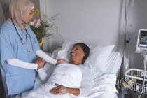 Front view of mixed race female doctor in hijab consoling mature mixed race female patient in the ward at hospital. — Stock Photo