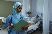 Vista frontal de la doctora de raza mixta con hijab mirando el informe médico mientras el paciente masculino caucásico duerme en la cama en la sala del hospital - foto de stock
