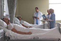 Vista lateral de diversos médicos que interactúan con el paciente masculino de raza mixta mientras que el paciente masculino caucásico duerme en la cama junto a ellos en la sala del hospital
. - foto de stock