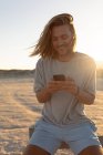 Vista frontal de hombre caucásico joven feliz usando el teléfono móvil en la playa - foto de stock
