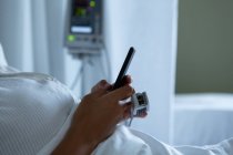 Partie médiane de la patiente utilisant un téléphone portable alors qu'elle était couchée dans le lit dans la salle d'hôpital — Photo de stock