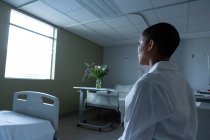 Vue latérale d'une patiente réfléchie de race mixte assise sur le lit dans la salle tout en regardant par la fenêtre à l'hôpital . — Photo de stock