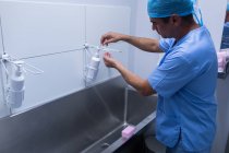 Vista lateral del cirujano caucásico de mediana edad lavándose las manos con jabón en el lavabo del hospital. Lleva bata quirúrgica y gorra. . - foto de stock