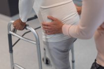 Bassa sezione di fisioterapista femminile che aiuta la paziente di razza mista a camminare con walker in ospedale — Foto stock