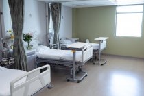 Modern hospital ward with empty beds, medical monitor, green curtains, cupboards, and flowers. — Stock Photo
