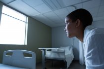 Vista lateral de la paciente femenina de raza mixta pensativa sentada en la cama mientras mira afuera a través de la ventana en la sala en el hospital — Stock Photo