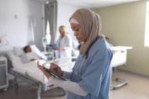 Вид збоку на жінку-лікаря змішаної раси в хіджабі, що пише в буфері обміну в лікарні. На задньому плані різні лікарі взаємодіють зі своїми пацієнтами . — стокове фото