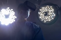 Visão de baixo ângulo de cirurgiã branca usando fone de ouvido de realidade virtual na sala de cirurgia do hospital. Luzes médicas estão no fundo . — Fotografia de Stock