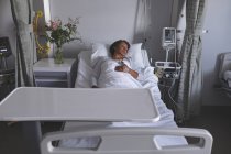 Vorderansicht einer reiferen Patientin mit gemischter Rasse, die mit einer Hand auf dem Bauch im Bett auf der Station im Krankenhaus schläft. Blumen, Monitor und Schrank stehen neben ihrem Bett. — Stockfoto