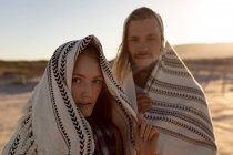 Vue de face du jeune couple caucasien enveloppé dans une couverture à la plage au coucher du soleil — Photo de stock