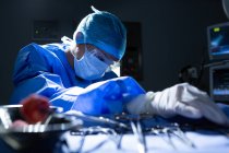 Vista frontal de una joven cirujana de raza mixta sosteniendo instrumentos quirúrgicos mientras realiza la operación en el quirófano del hospital - foto de stock